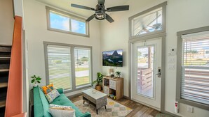 A bright living room with a teal sofa, a coffee table, and a ceiling fan. There are large windows with blinds, a TV mounted on the wall, and a wooden cabinet with a plant.