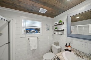 A bathroom with a toilet, sink, and window with blinds. There are shelves with towels and a mirror above the sink.