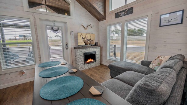 Ein gemütliches Wohnzimmer mit Kamin, einem grauen Sofa und einem großen Fenster mit Jalousien.