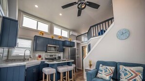 A modern kitchen with dark blue cabinets, a central island with stools, a microwave, and a stove. There is a staircase leading to an upper level with a clock on the wall.
