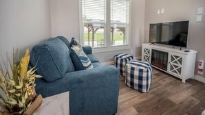 A living room with a blue sofa, ottomans, a TV on a white cabinet, and a window with blinds.