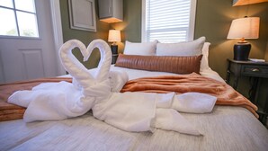 A neatly made bed with a heart-shaped towel arrangement, two bedside lamps, and a window with blinds. - Rinconcito Magico - 200 (Fredericksburg)