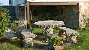 A stone outdoor seating area with a round table and benches, surrounded by a stone wall and greenery.