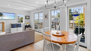 Una habitación amplia con una mesa de comedor de madera, sillas blancas, un sofá gris y un ventanal con vistas al exterior.