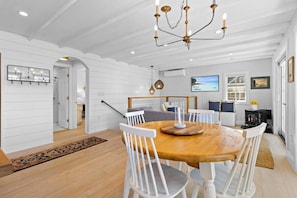 A dining area with a wooden table and white chairs, a chandelier, and a television in the background.