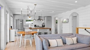 Una cocina moderna con muebles blancos, un comedor con mesa de madera y sillas blancas, y un sofá gris con cojines.