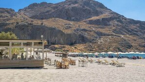 A beach with a rocky mountain backdrop, a wooden structure with tables and chairs, and umbrellas on the sand. - Villa w 2 pools, BBQ, Sauna,250m to Plakias Beach (Μυρθιανος Πλακιάς)