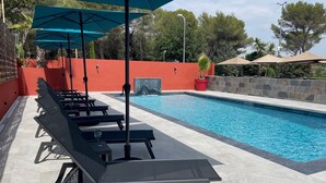 Zona junto a la piscina con sillas de descanso y sombrillas, un muro rojo y una piscina azul cristalina.