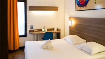 A hotel room with a bed, a desk, and a chair. There is a window with curtains, a painting on the wall, and a headboard.