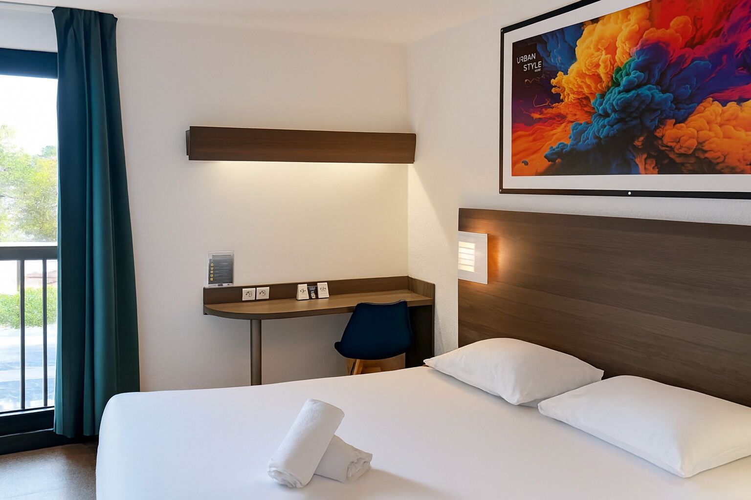 Hotelkamer met een bed, een bureau en een stoel. Er is een raam met gordijnen, een schilderij aan de muur en een bord met pictogrammen.