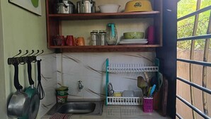 Una cocina pequeña con un lavabo, una ventana con barrotes, una estantería con utensilios de cocina y un cuadro de frutas en la pared.
