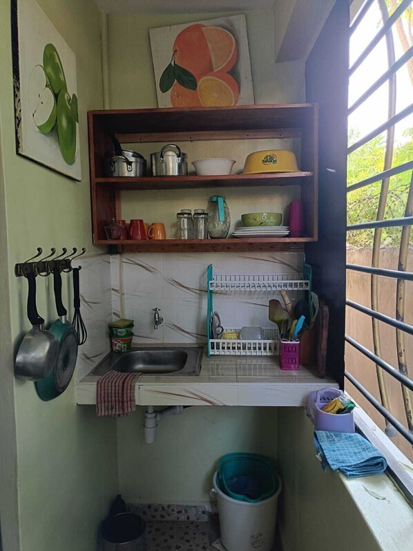 A small kitchen with a sink, a window with bars, a shelf with kitchen utensils, and a painting of fruit on the wall.