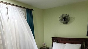Un dormitorio con una cama, dos sillones tapizados, una mesita de noche con un florero y un ventilador de pared.