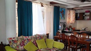 Una sala de estar con un sofá floral, una mesa de comedor de madera, sillas y una zona de cocina con estufa y utensilios.