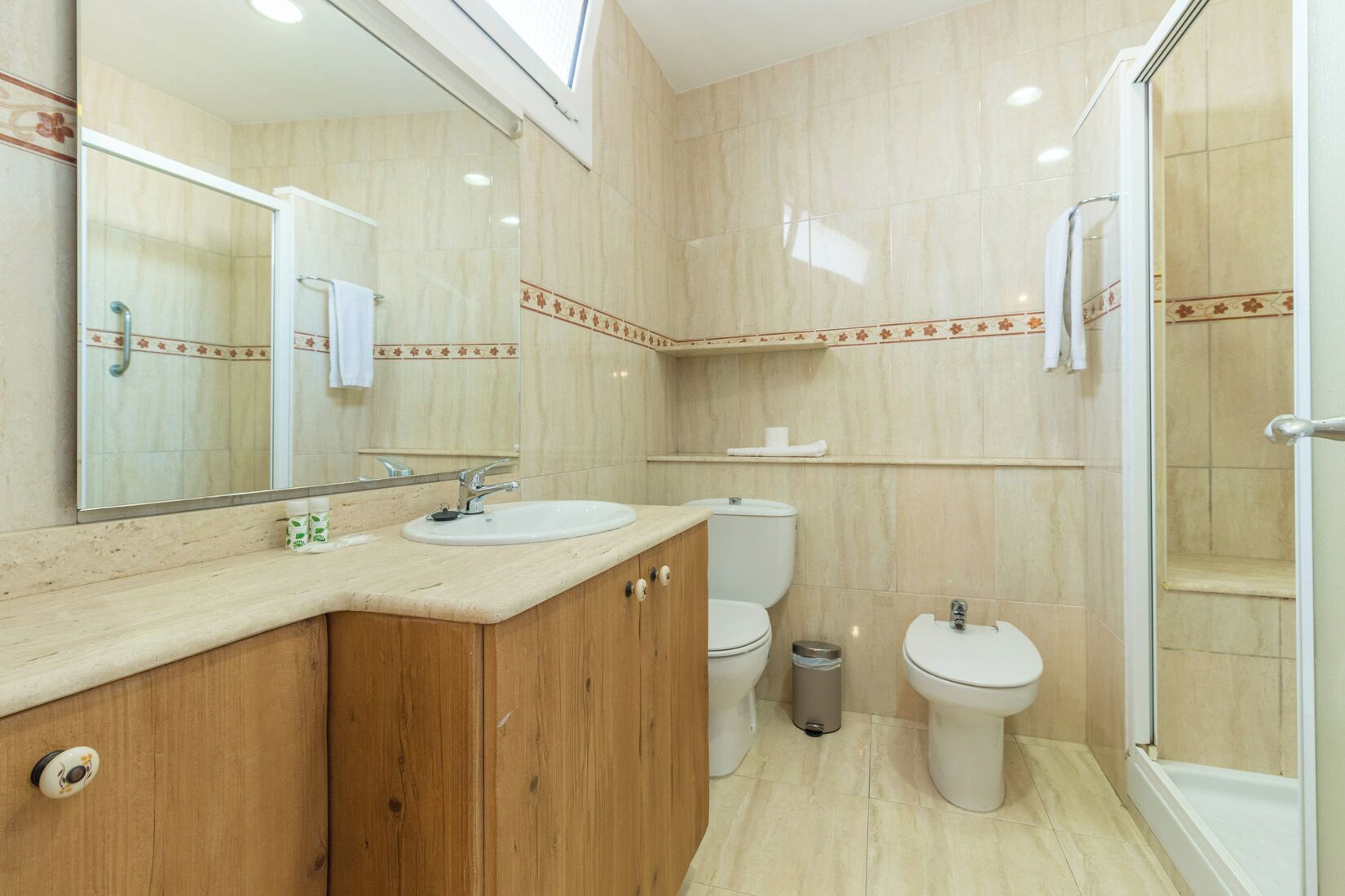 Baño con tocador de madera, dos inodoros, un lavamanos, un espejo y una ducha.