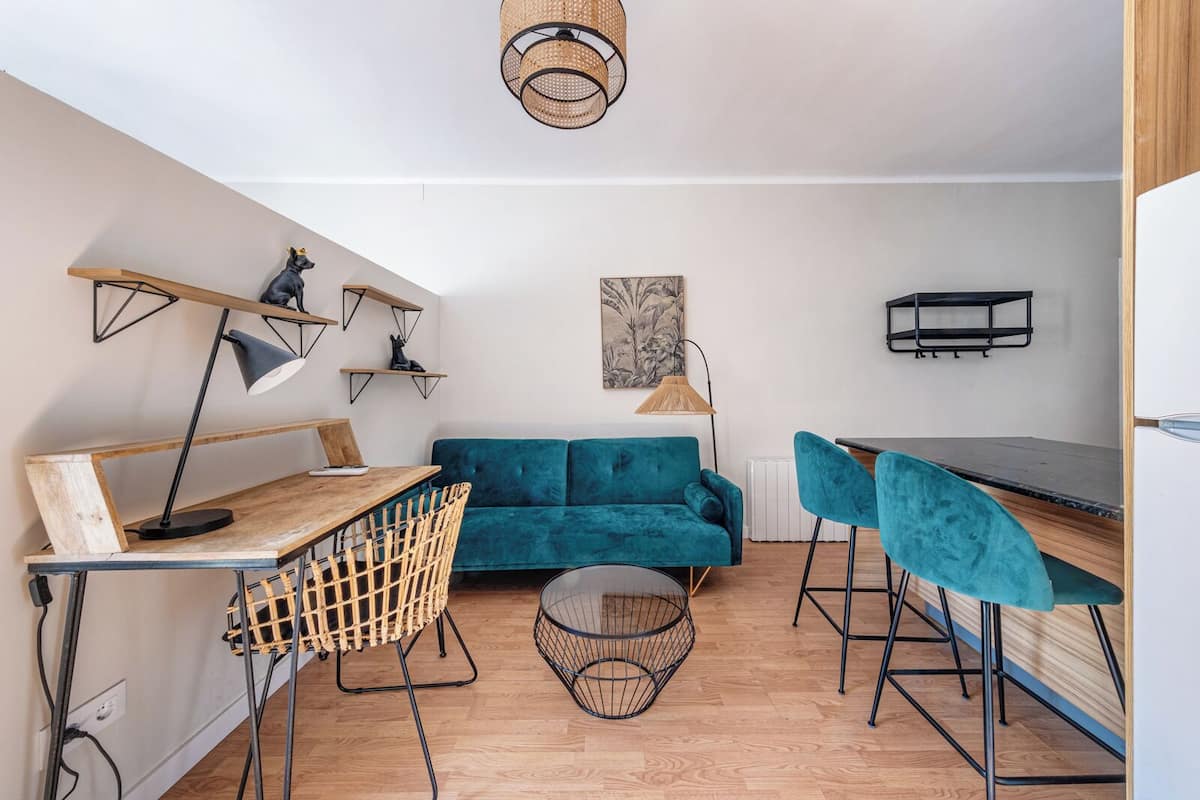 A compact living space with a teal sofa, a wooden dining table, and a small kitchen area.