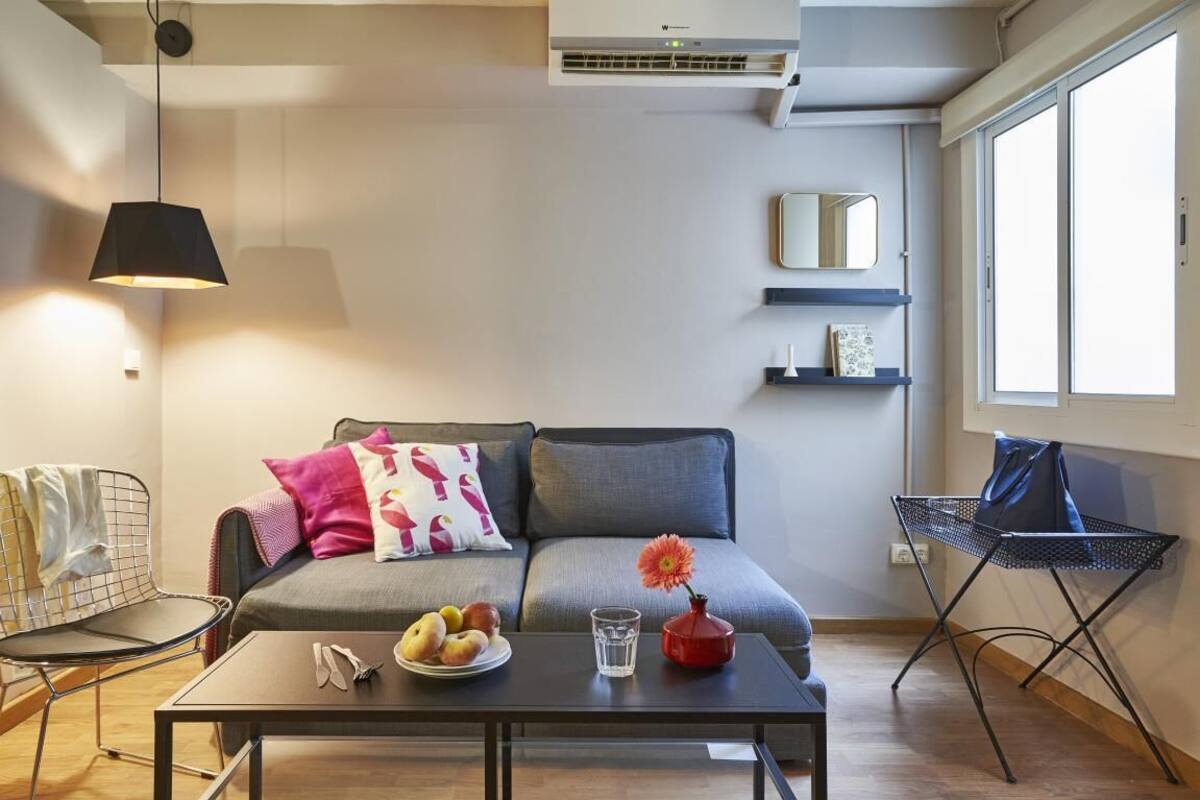 A modern living room with a sofa, a coffee table with fruits and a vase, a hanging lamp, and an air conditioning unit.