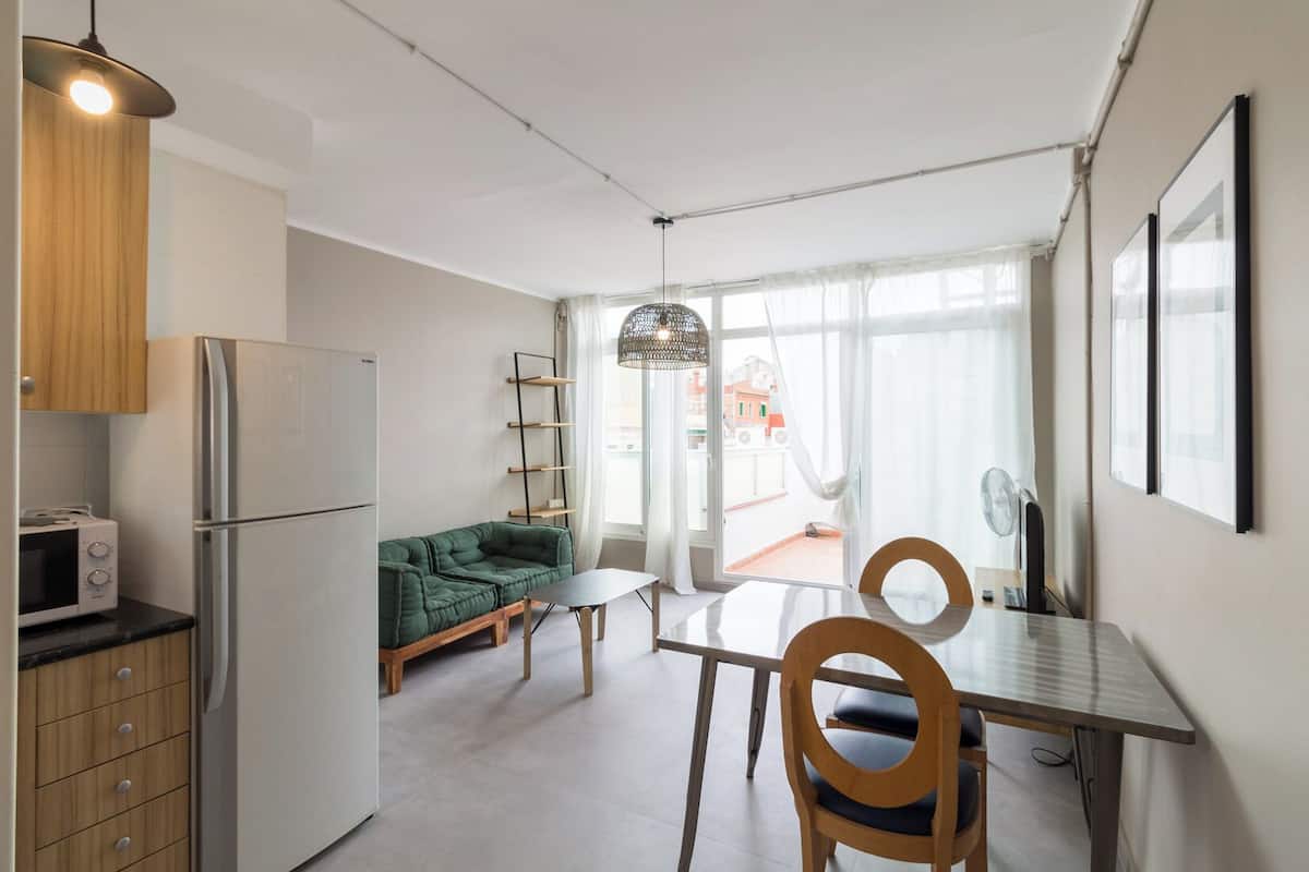 A compact kitchen with a refrigerator, microwave, and wooden cabinets. A green sofa and a dining table with chairs are in the living area, which has large windows and a view of buildings outside.