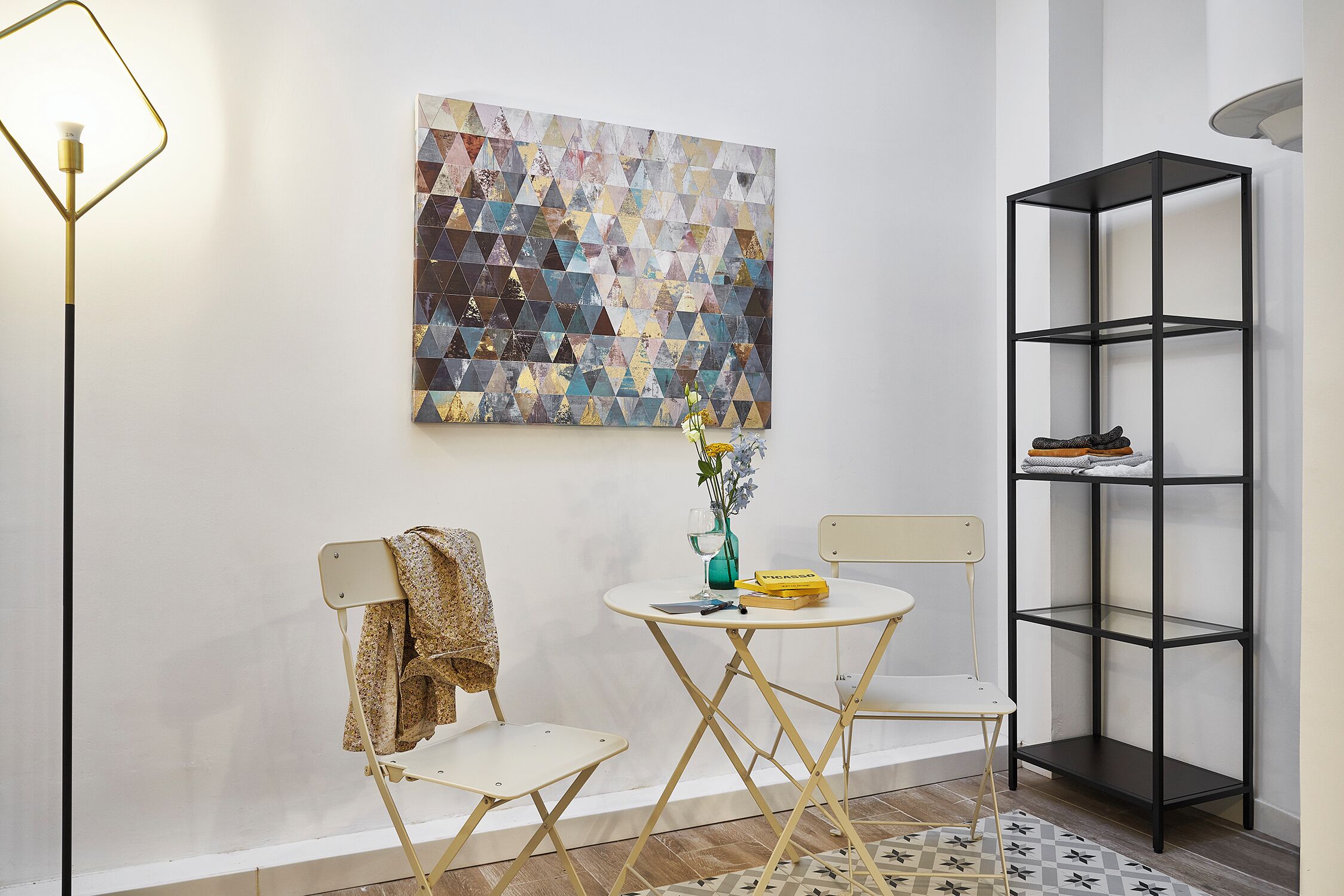 Una habitación con un cuadro de pared con motivos geométricos, un estante de metal negro, una pequeña mesa redonda con un jarrón y una silla, y una silla plegable con una chaqueta.