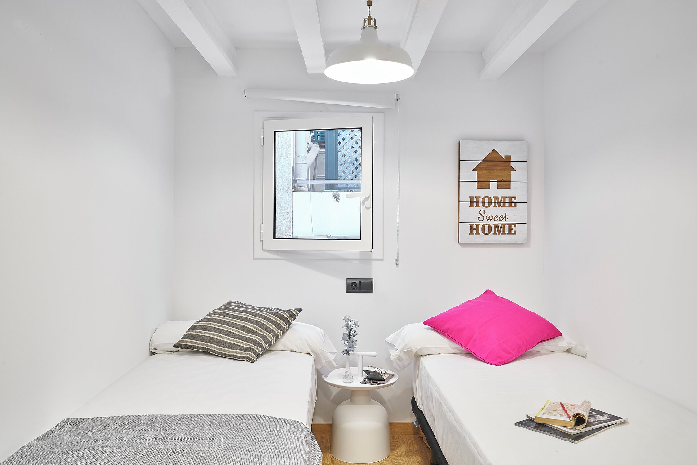 A small, clean room with two beds, a window, a wall-mounted light, and a "HOME Sweet HOME" sign.