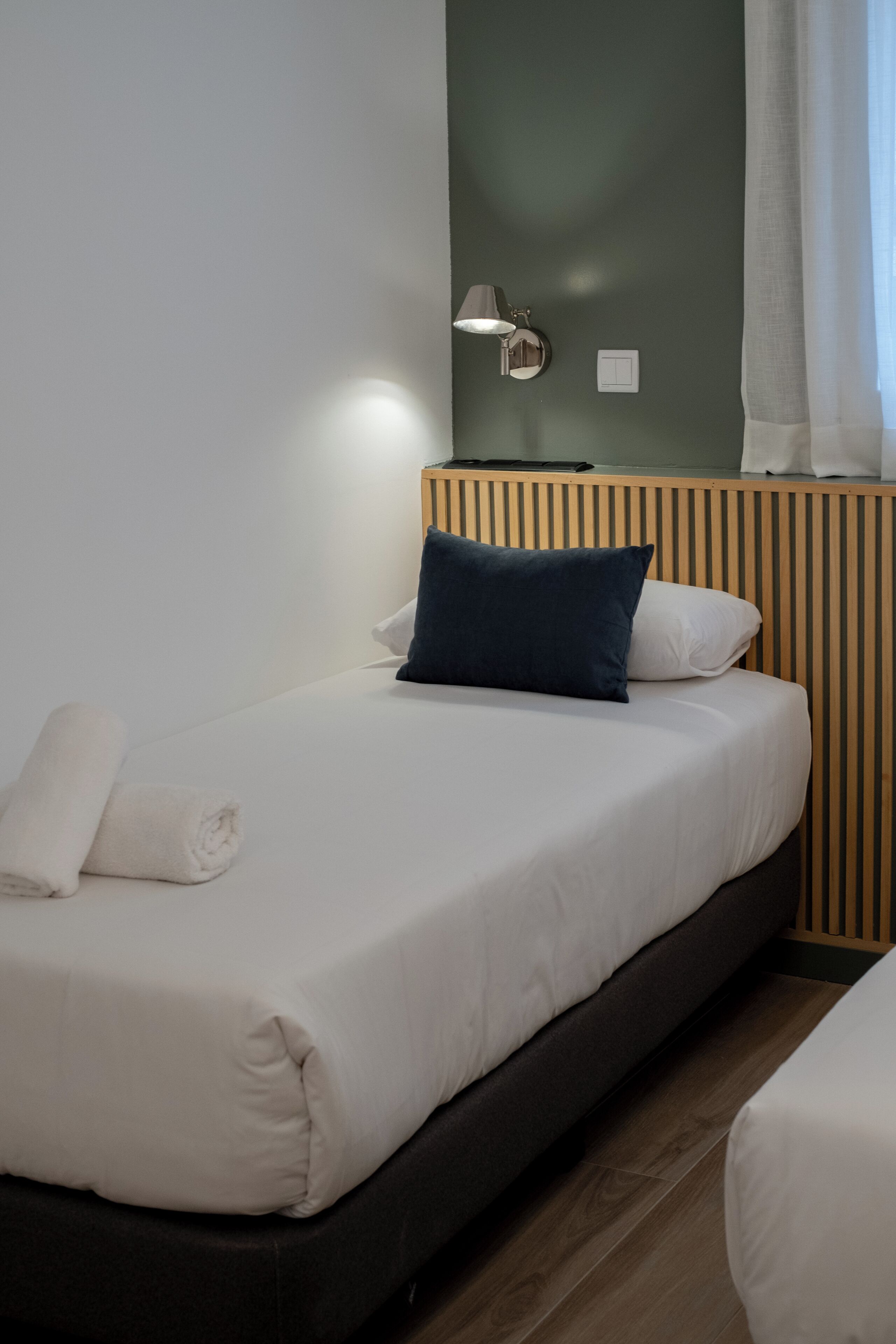 Un lit simple avec un matelas blanc et un oreiller bleu foncé, une tête de lit en bois, une lampe fixée au mur et une fenêtre avec des rideaux légers.