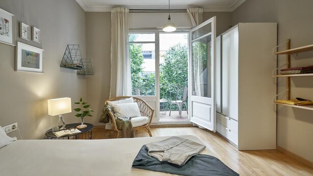 Hotelkamer met een bed, een stoel, een klein tafeltje met een lamp, een boekenkast en een schuifdeur naar een balkon.