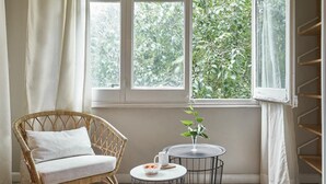 Een kamer met een rieten stoel, een klein rond tafeltje met een plant en een raam met uitzicht op groen.