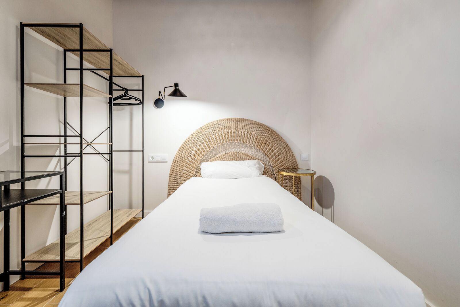 Quarto de hotel minimalista com uma cama, um guarda-roupa de madeira, uma pequena mesa redonda e uma luminária fixada na parede.