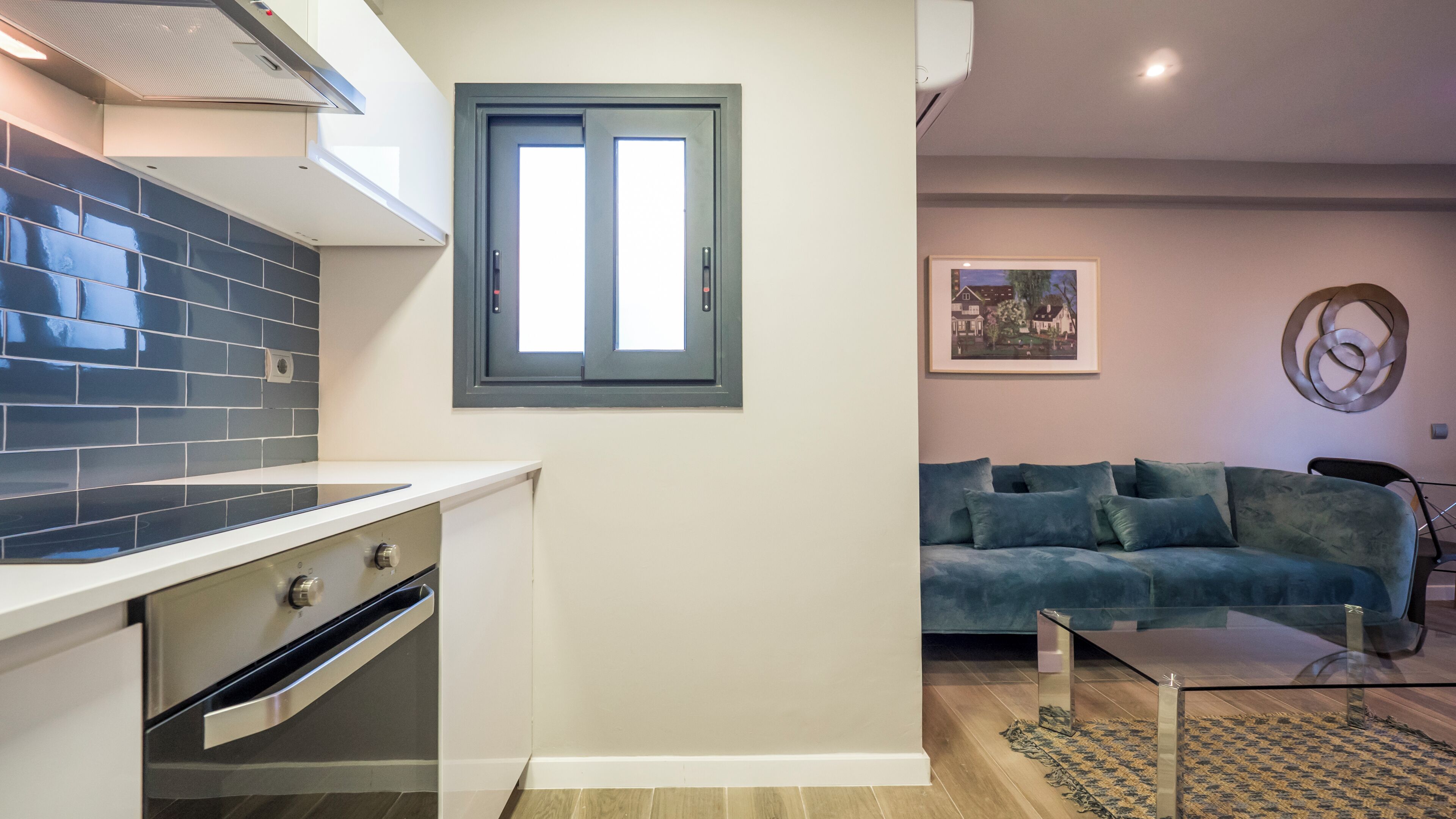 Una cocina moderna con horno empotrado, ventana con vistas, sofá azul y mesa de centro de vidrio.