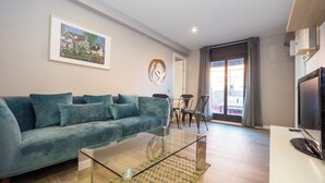 Un salon avec un canapé de couleur turquoise, une table basse en verre, un tableau au mur, un coin repas avec des chaises et un téléviseur posé sur un meuble.