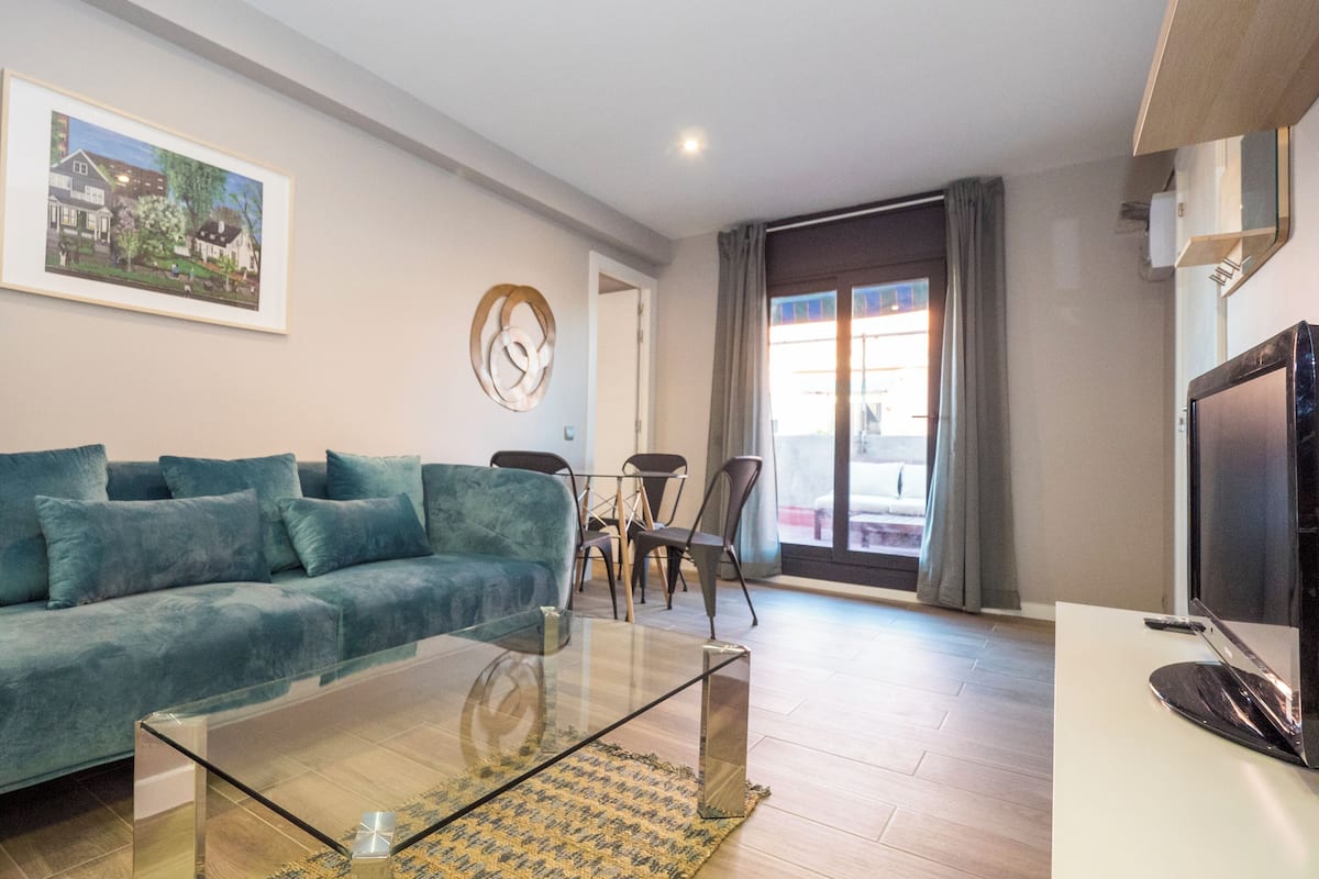 Ein Wohnzimmer mit einem petrolfarbenen Sofa, einem Glastisch, einem Gemälde an der Wand, einem Essbereich mit Stühlen und einem Fernseher auf einem Standfuß.