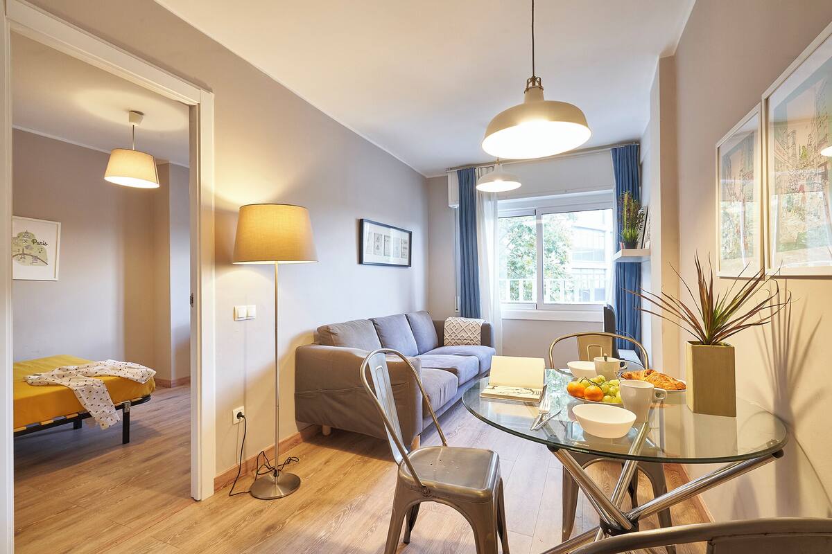 Ein modernes Wohnzimmer mit einer Couch, einem Glastisch mit Stühlen, einer Lampe und einem Fenster mit Vorhängen.