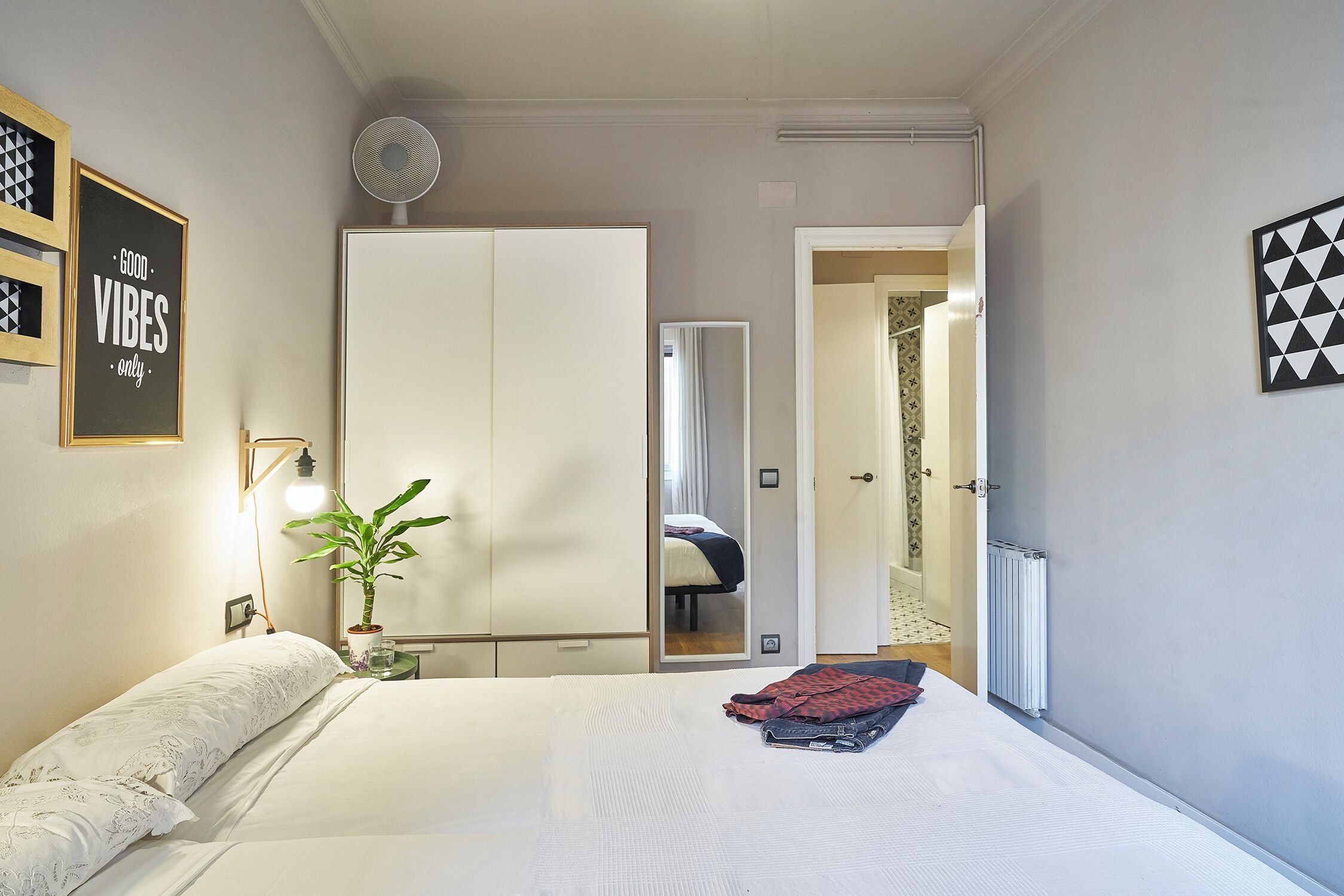 Un dormitorio con cama, mesita de noche con lámpara, espejo, radiador y puerta que da a otra habitación.