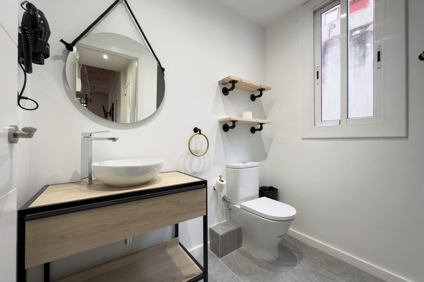 Un baño moderno con un lavamanos blanco, un inodoro y un espejo. Hay una ventana con vistas al exterior.