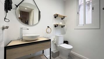 Un baño moderno con un lavamanos blanco, un inodoro y un espejo. Hay una ventana con vistas al exterior.