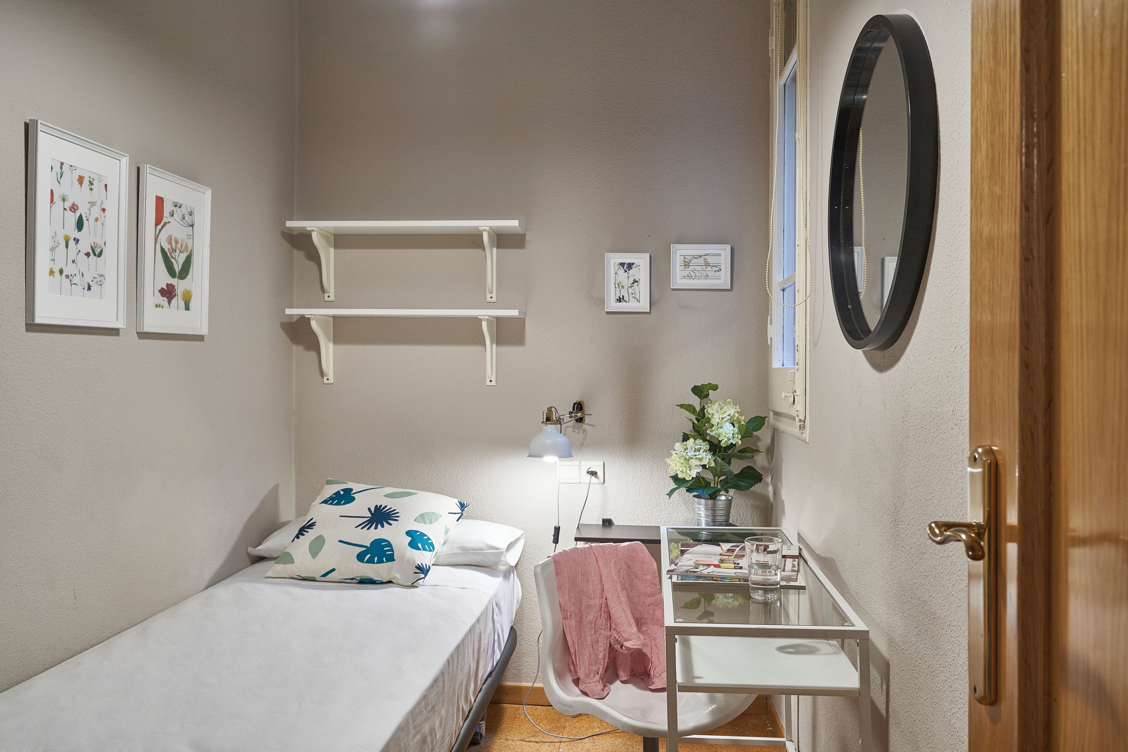 A small, neatly arranged bedroom with a single bed, a bedside table, a mirror, and wall-mounted shelves.
