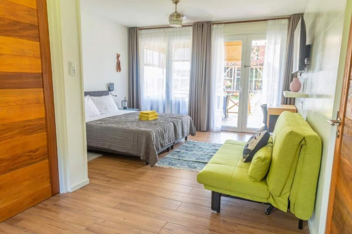 Un dormitorio con una cama, un sofá verde, una mesita pequeña y un ventanal corredero que da a un espacio exterior.