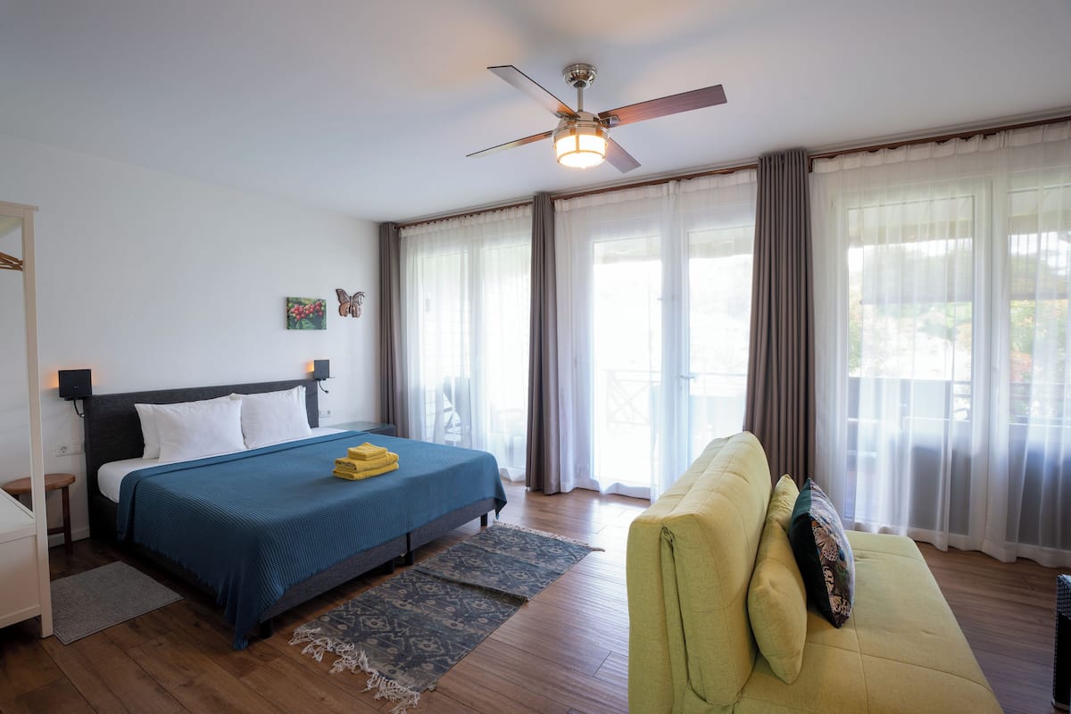Un dormitorio con cama, sofá verde, ventilador de techo y ventanas grandes con cortinas.
