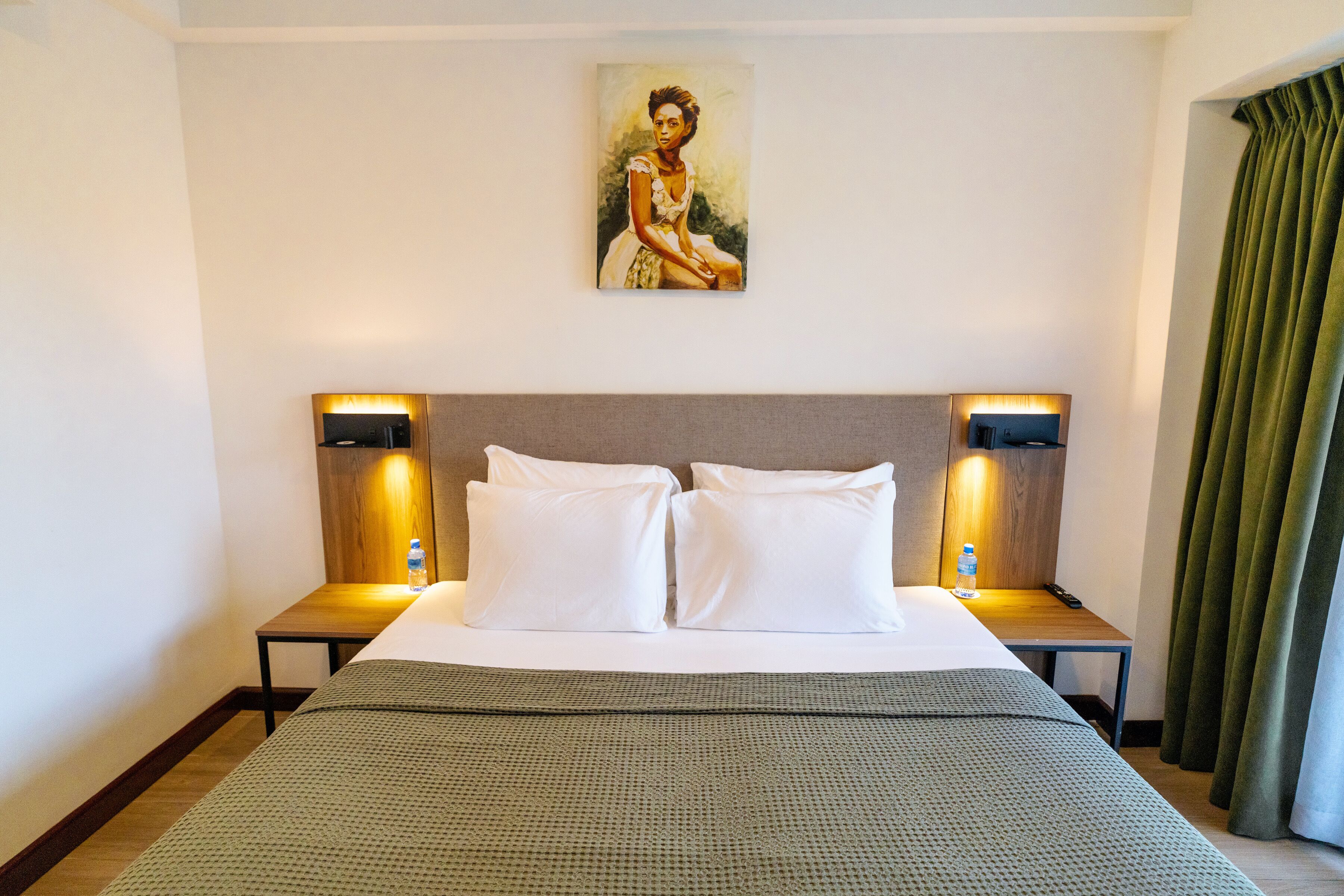 Quarto de hotel com uma cama grande, duas mesas de cabeceira com abajures, um quadro na parede e uma cortina verde.