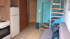 A compact kitchen with a refrigerator, stove, and wooden cabinets. A staircase leads to a bedroom area with a sofa and pillows.
