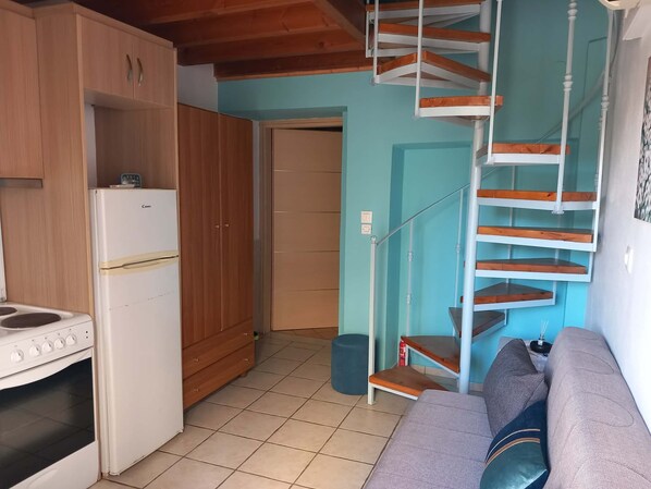 Ett kompakt kök med kylskåp, spis och träskåp. En trappa leder upp till ett sovområde med en soffa och kuddar.