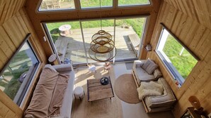 A high-ceilinged wooden cabin with a loft bedroom, a cozy living area with a sofa and a hanging chair, and a view of a green landscape through large windows. - Quiet lodge in Wissant, in the forest and 500m from the beach.  Ideal for a couple (Wissant)