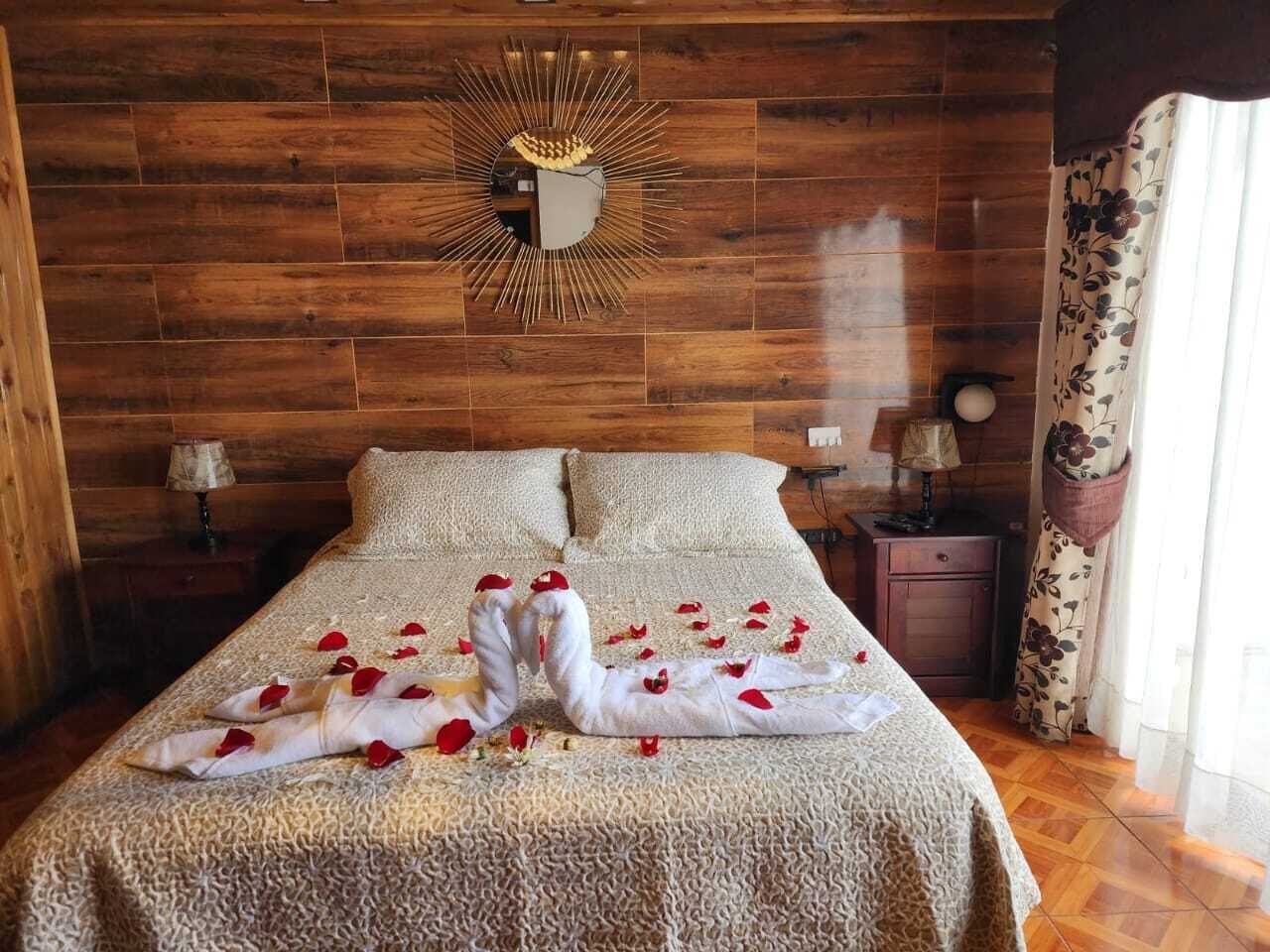 A bed with rose petals and a towel sculpture, wooden panel walls, and a nightstand with a lamp.