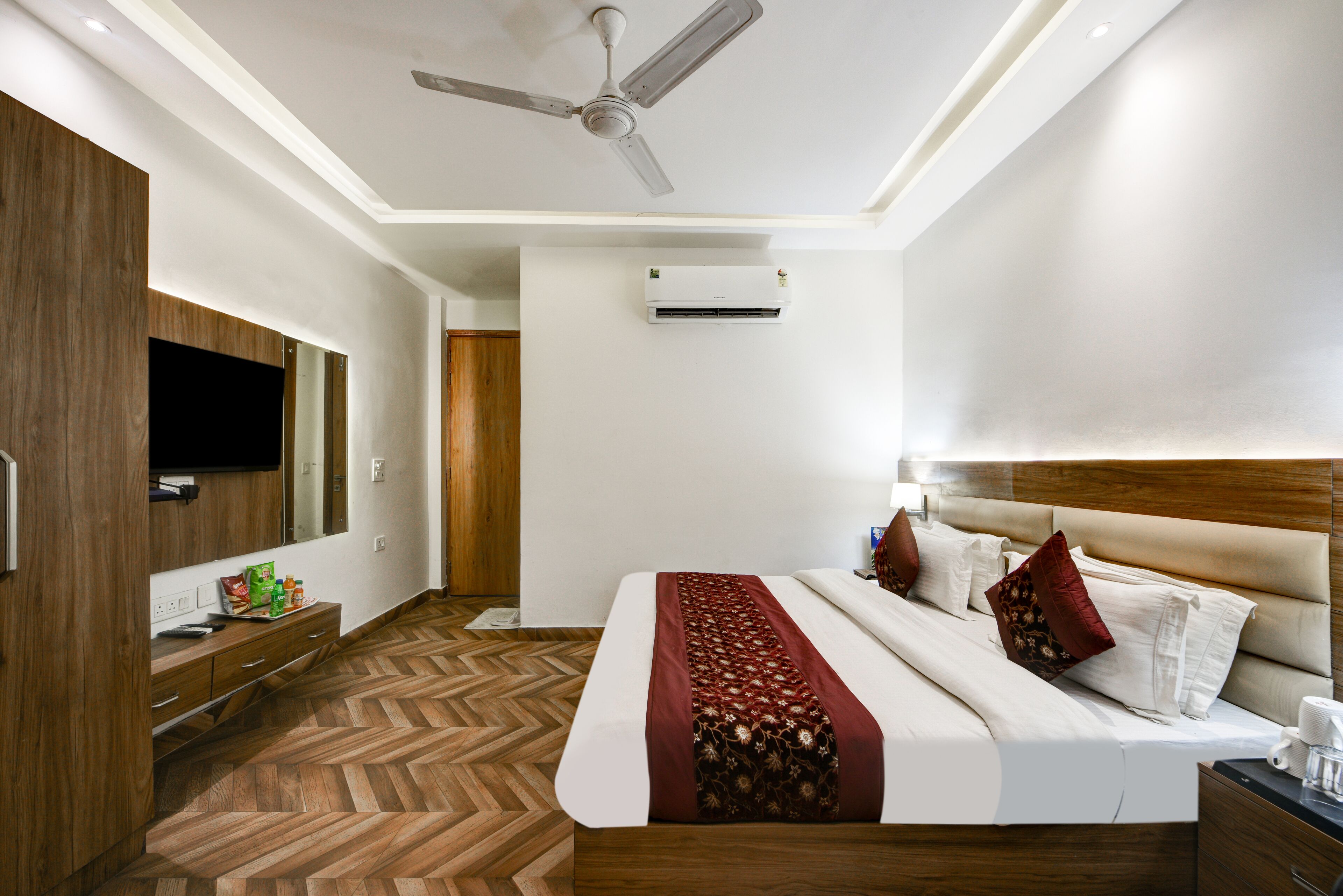 A hotel room with a bed, wooden wardrobe, TV, air conditioning unit, and a ceiling fan.