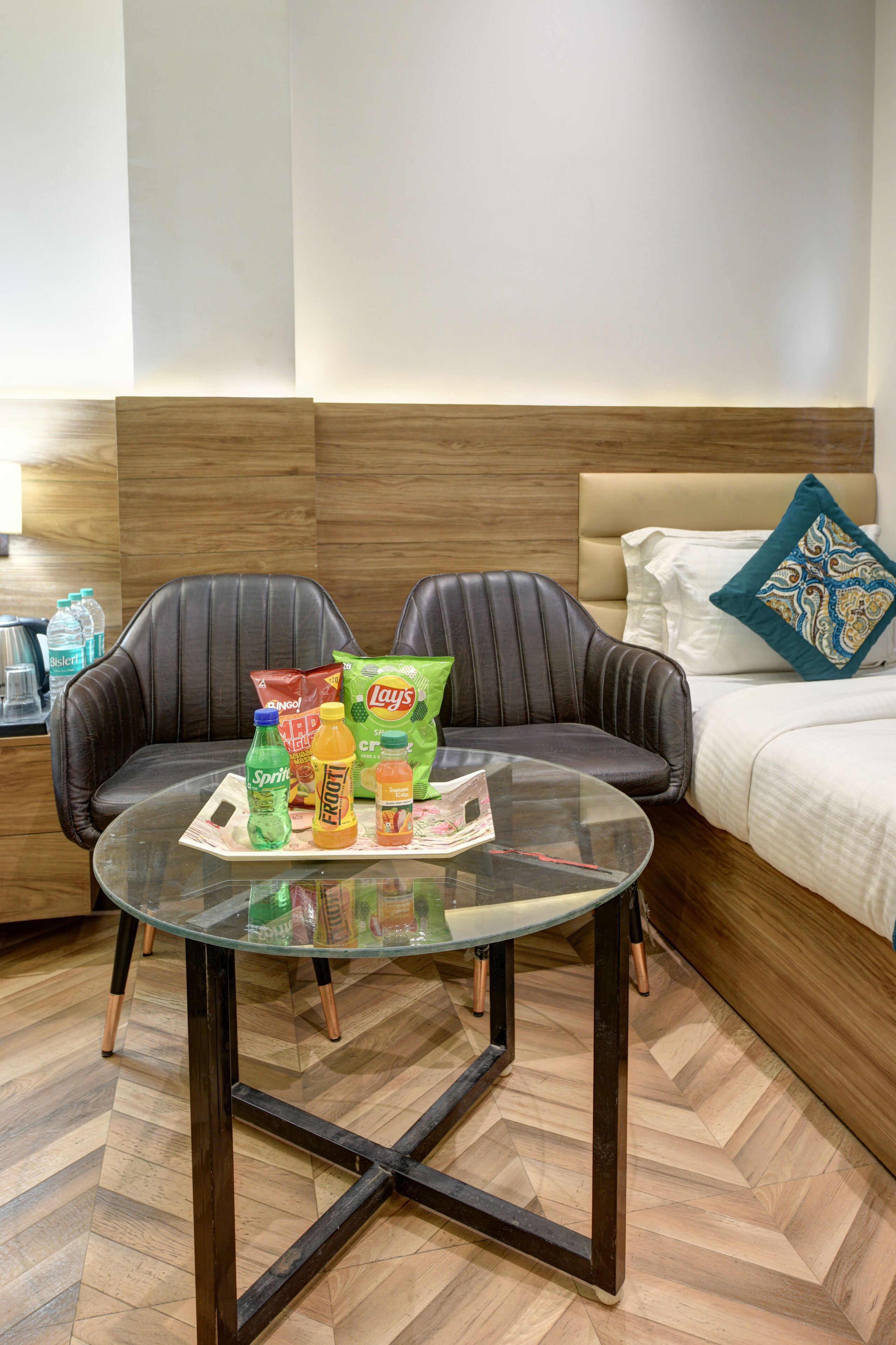 Une chambre d’hôtel avec une table à plateau en verre, deux fauteuils en cuir, un lit avec un oreiller blanc et bleu, et des en-cas sur la table.