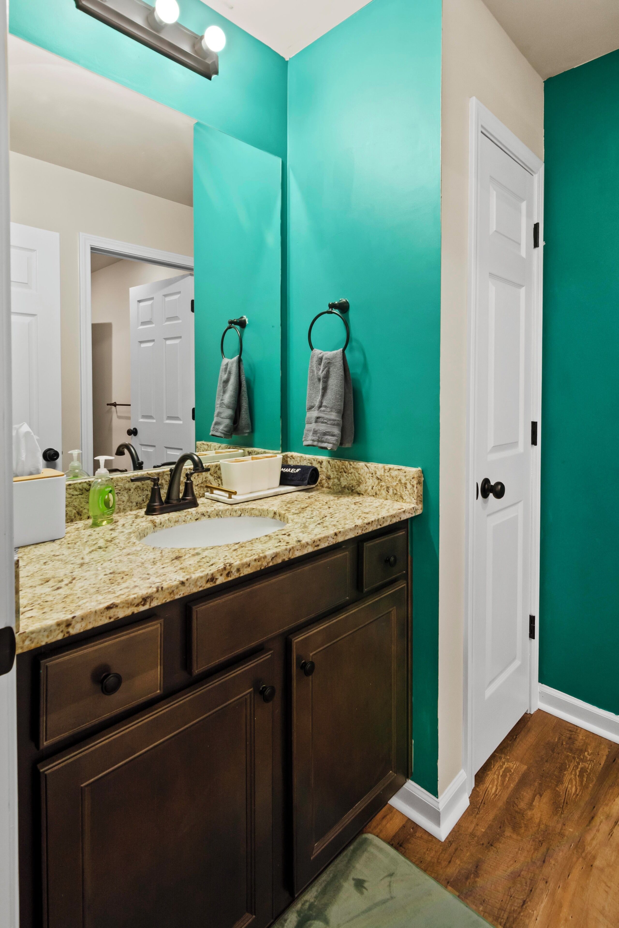 Une salle de bain avec un lavabo sur un plan de travail en granit, des meubles en bois foncé, des murs de couleur turquoise et une porte blanche.