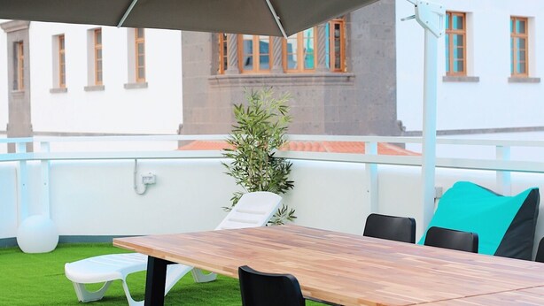 Un área de estar al aire libre con una mesa de madera, sillas negras, un paraguas grande y una planta en maceta.