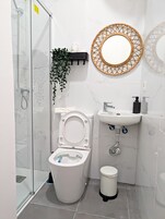 Un baño moderno con cabina de ducha de vidrio, inodoro, lavabo con espejo y un pequeño cubo de basura.