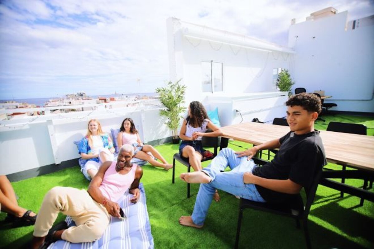 A rooftop terrace with a view of the city, a wooden table, and several people sitting and relaxing.