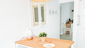 Un comedor luminoso y blanco con una mesa de madera, dos sillas blancas, una lámpara colgante de mimbre y un ventanal con marcos blancos.